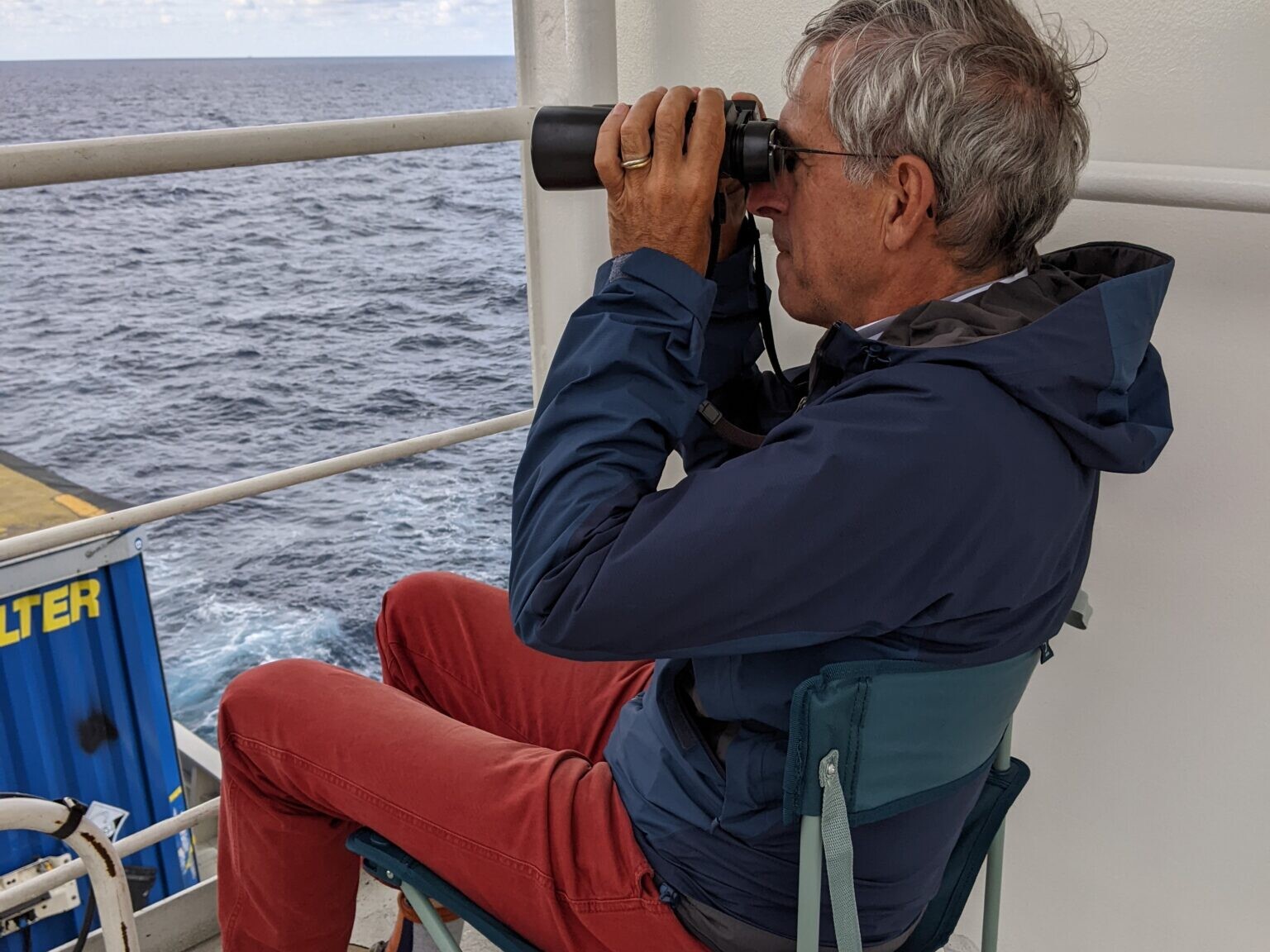 man looks through binoculars with view of sea and containers
