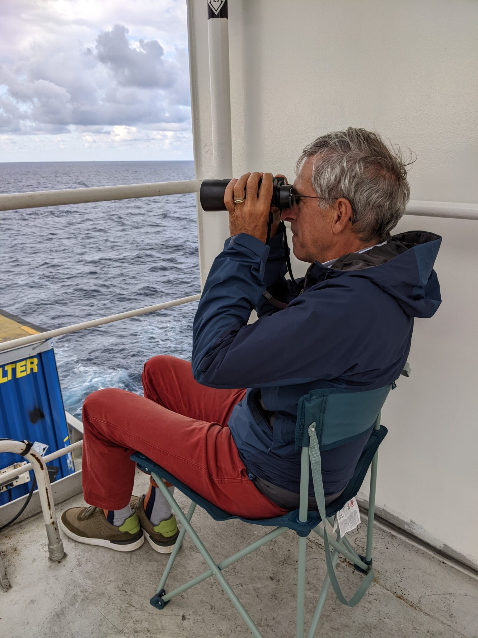 man looks through binoculars with view of sea and containers